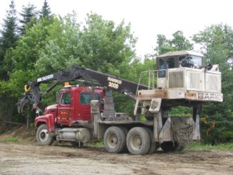 A three-axle truck with a loader mounted to the far back of the truck is parked at the side of a landing. The grapple and boom are extended and rest above the truck cab.
