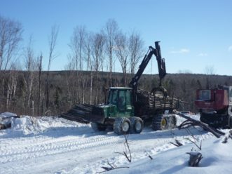 An eight-wheeled forwarder fully loaded with a knuckle-boom on top of the logs in northern Maine. Snow is on the ground and surrounding the forwarder.