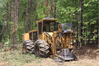 A four-wheeled feller-buncher parked in front of a pine plantation with the feller-buncher head showing the sawdisk