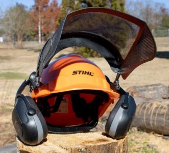 A picture of a hardhat safety system with earmuffs and a face shield displayed on top of a wooden log.