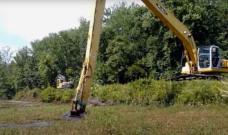 An excavator sits on the upland bank and uses the bucket to scoop sediment out of the pond.